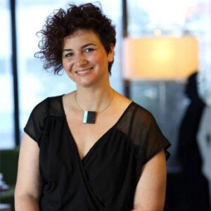 A young person with curly hair smiles at the camera while sitting in front of a laptop against a dark background.