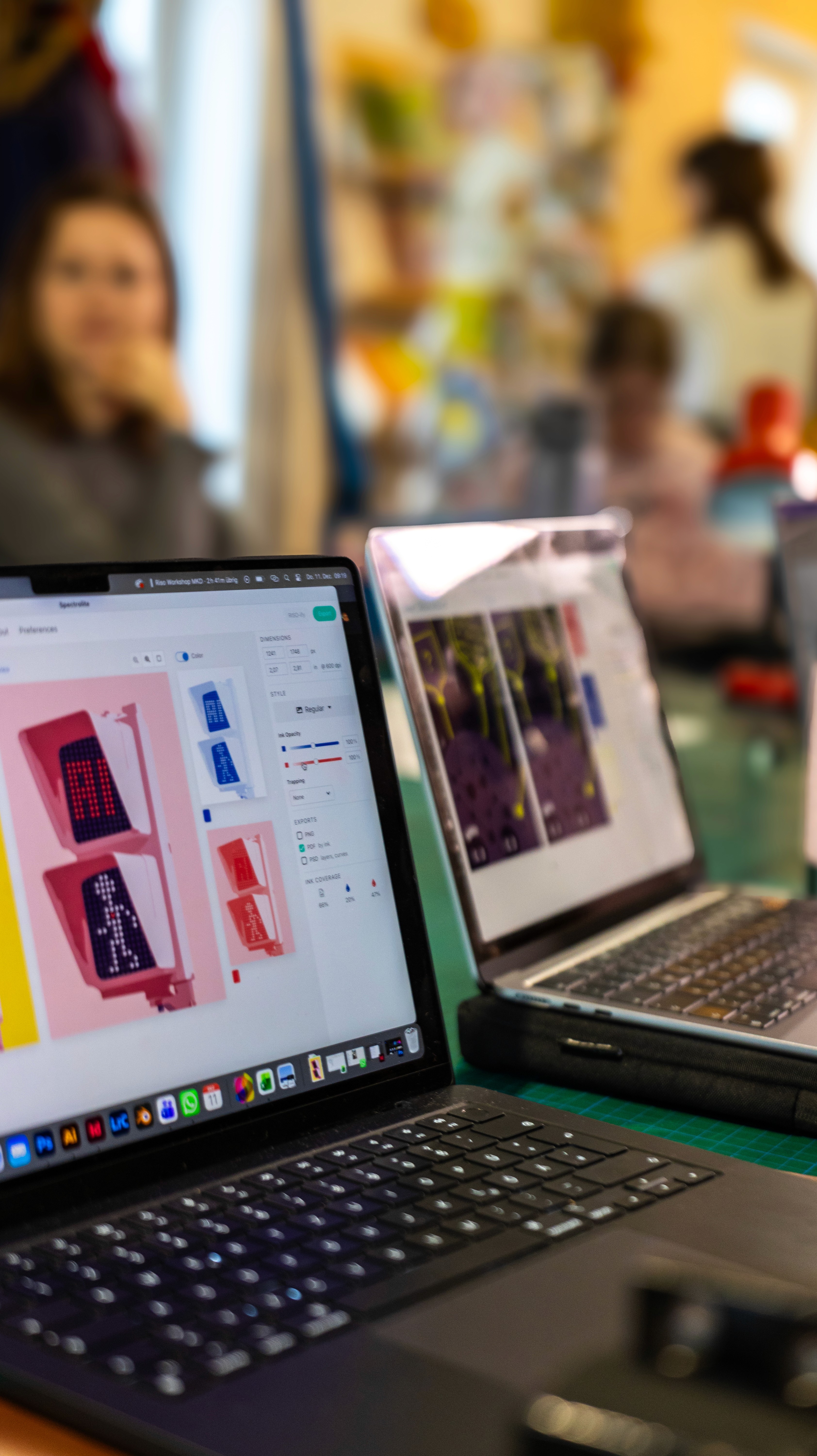 A laptop with colorful designs displayed on the screen, with people chatting and working in the background.