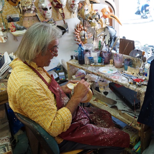 Una persona con una maglietta gialla e un grembiule rosso realizza una maschera in un laboratorio pieno di attrezzi, colori e maschere.