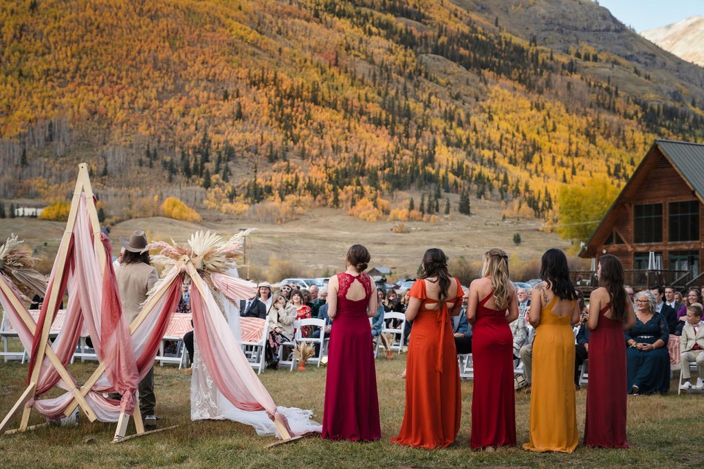 A line of bridesmaids in vibrant autumnal shades of red, orange, and gold stand before a dramatic mountain backdrop during a ceremony captured by a Silverton Colorado wedding photographer.