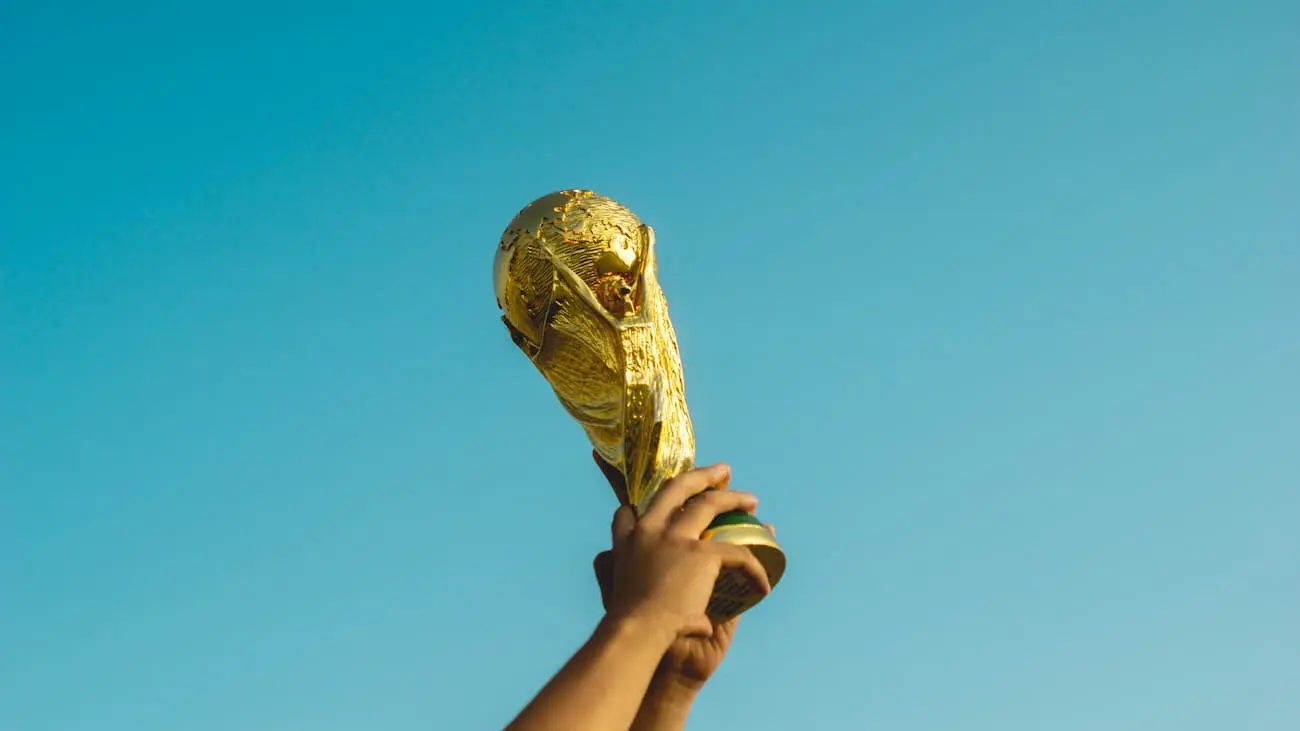 Capturing the quintessence of football glory, the image showcases the iconic FIFA World Cup Trophy held high, basking in the sunlight. The golden trophy stands out against the serene blue backdrop of the sky, symbolising the pinnacle of international football success. This moment frozen in time represents the ultimate achievement every football player, coach, and fan dreams of, reflecting the joy and pride of being crowned world champions.