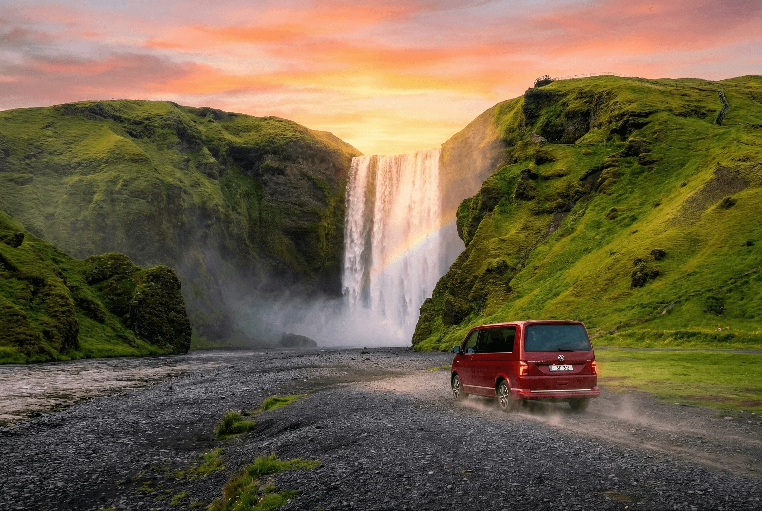 A red camper van driving on a gravel path towards a large waterfall with a visible rainbow.