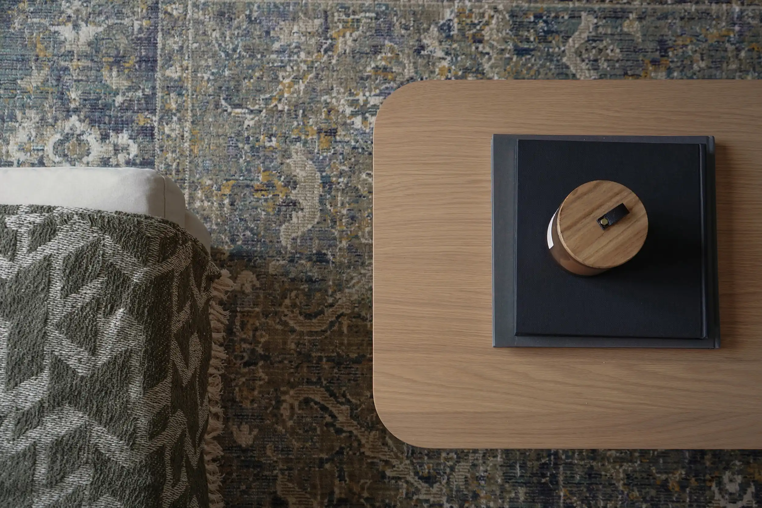 Styled living room detail with an oak side table, wooden decorative accent, charcoal tray, olive knit throw blanket, and a vintage Persian-style area rug — interior design by The Simone Co. in Fort McMurray