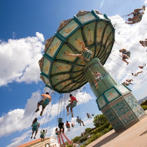 Personas disfrutando de un gran columpio en un parque de atracciones bajo un cielo azul con nubes.