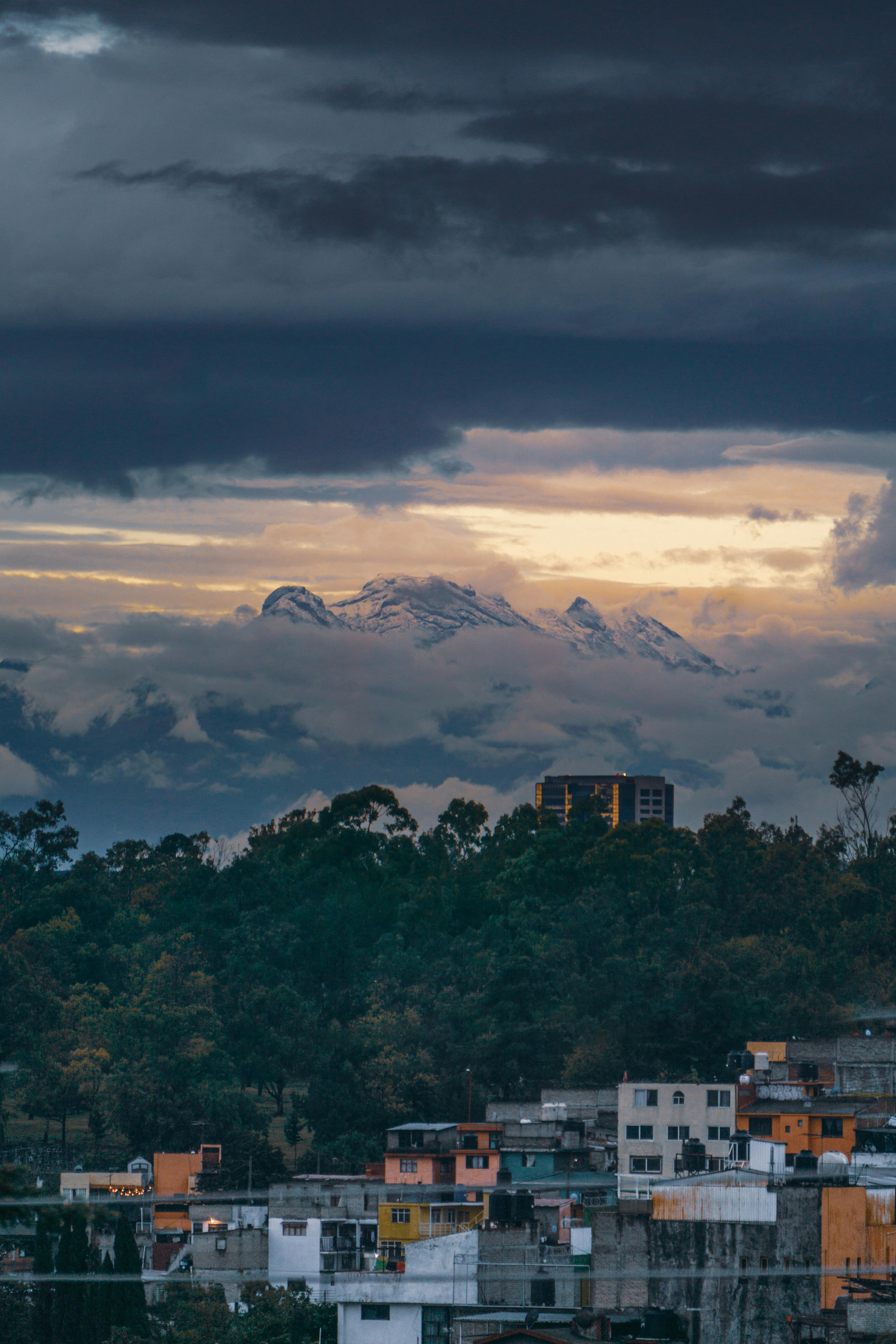 A view of a city neighborhood at twilight with a majestic, snow-capped mountain range visible in the distance through a gap in the dark, heavy clouds.