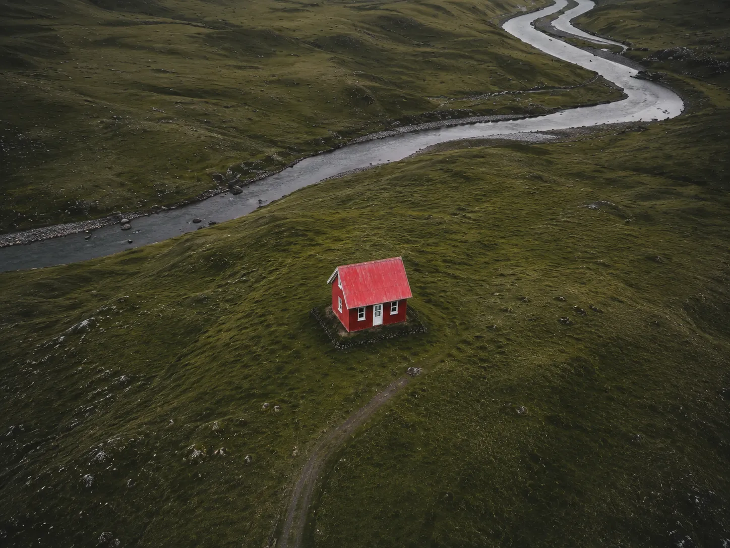 Drone view of a red mountain chalet surrounded by green hills and a winding river in a remote landscape.