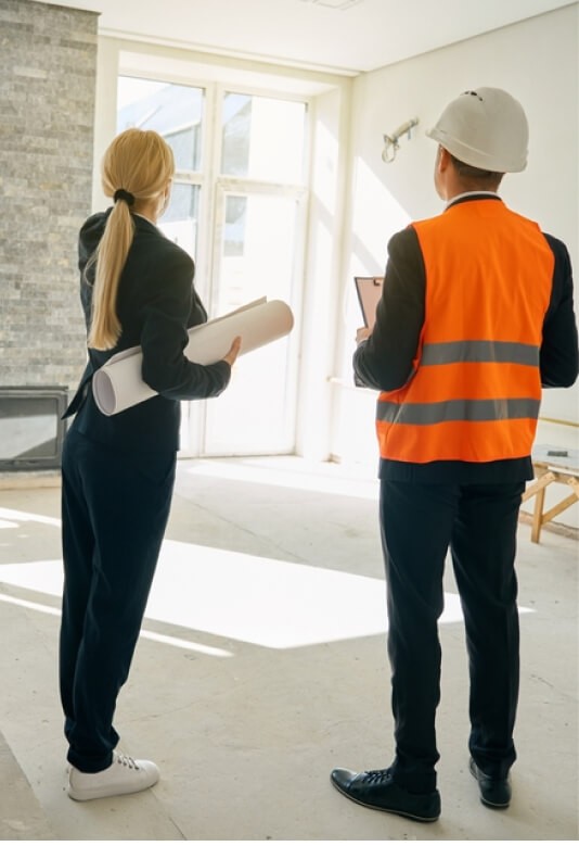 Two construction professionals discuss plans in an unfinished room, one in a safety vest and hard hat.