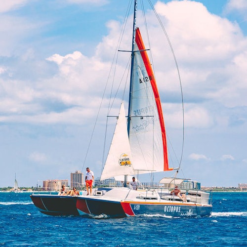 Des personnes se détendent sur un catamaran nommé "OCTOPUS" naviguant sur un océan bleu sous un ciel partiellement nuageux, avec des immeubles lointains visibles.