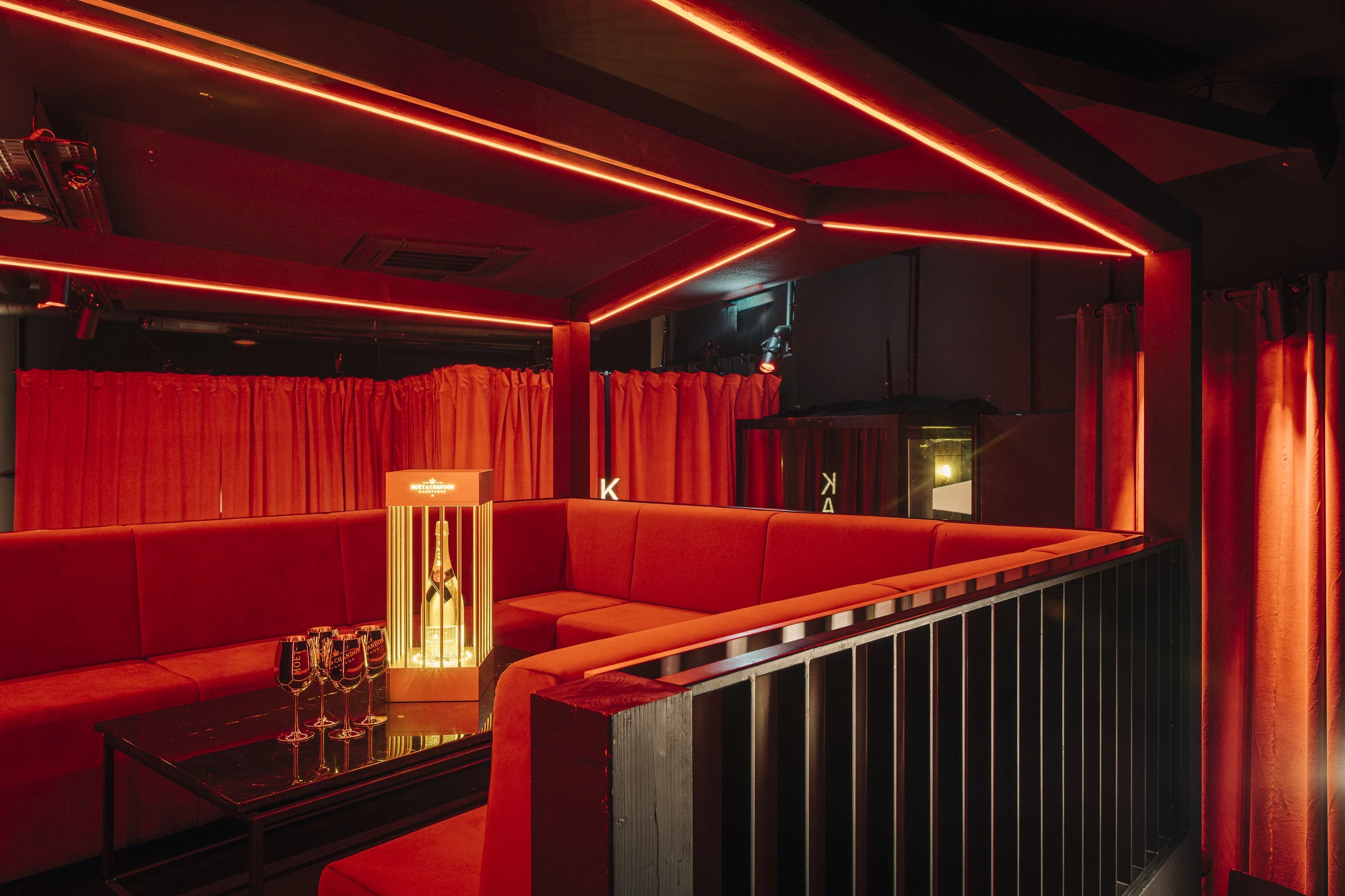 Red-lit VIP lounge at NATE CLUB with a U-shaped velvet sofa, red curtains, neon ceiling lines and an illuminated champagne display on the table.