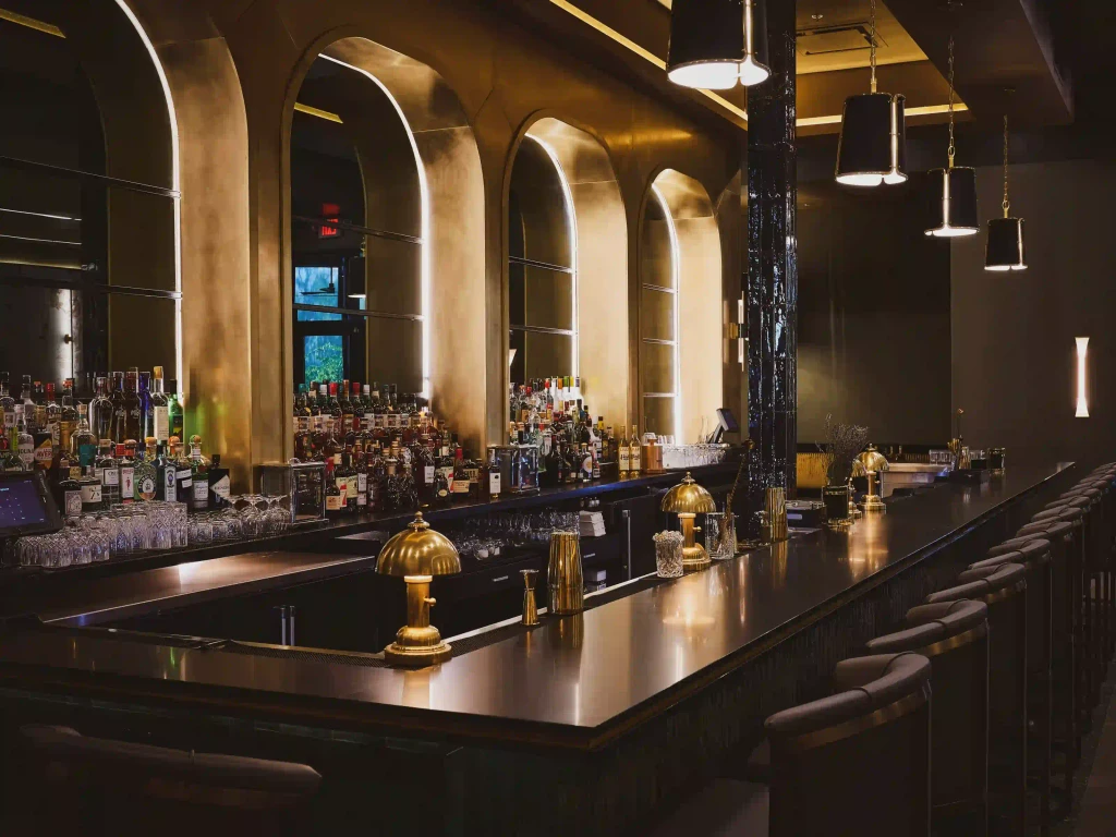 A long, elegant bar with a dark wood top, gold accents, and arched mirrors. Bottles of liquor line the back.