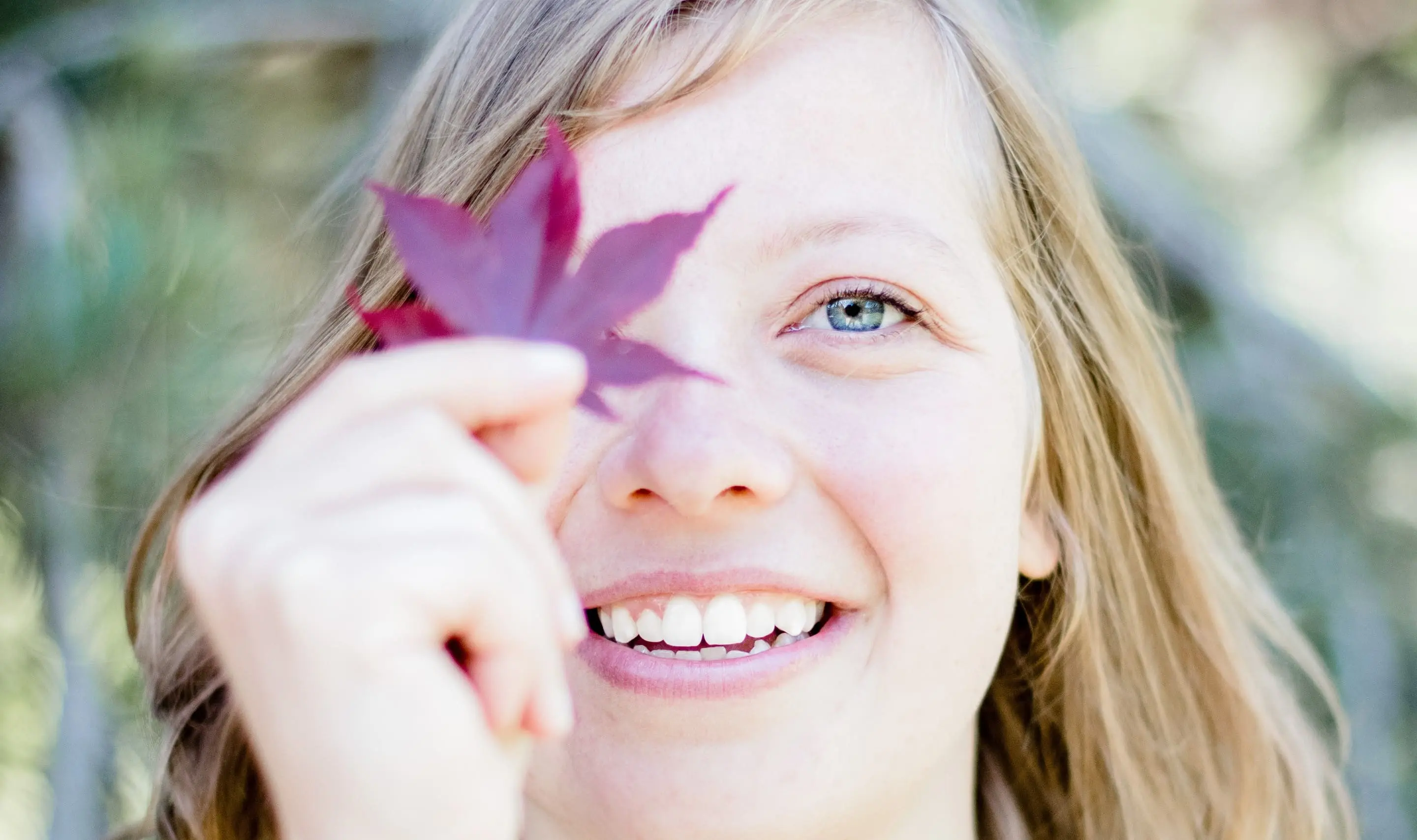 Close-up of a woman looking at the camera, smiling warmly, and holding a leaf in front of her right eye.