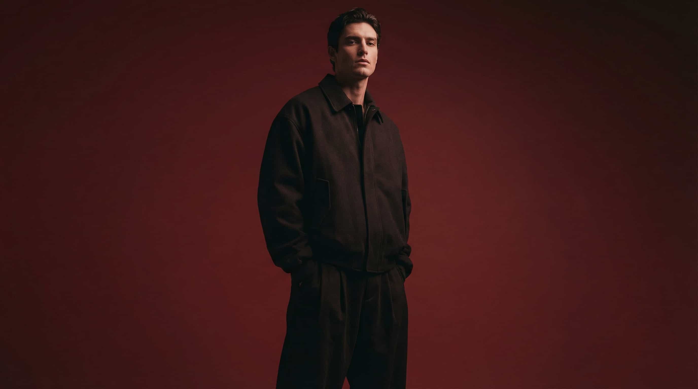 Male fashion model in dark clothing against a red studio background.
