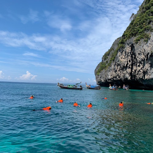 People in orange life jackets snorkeling near boats and rocky cliffs in clear blue water on a sunny day.