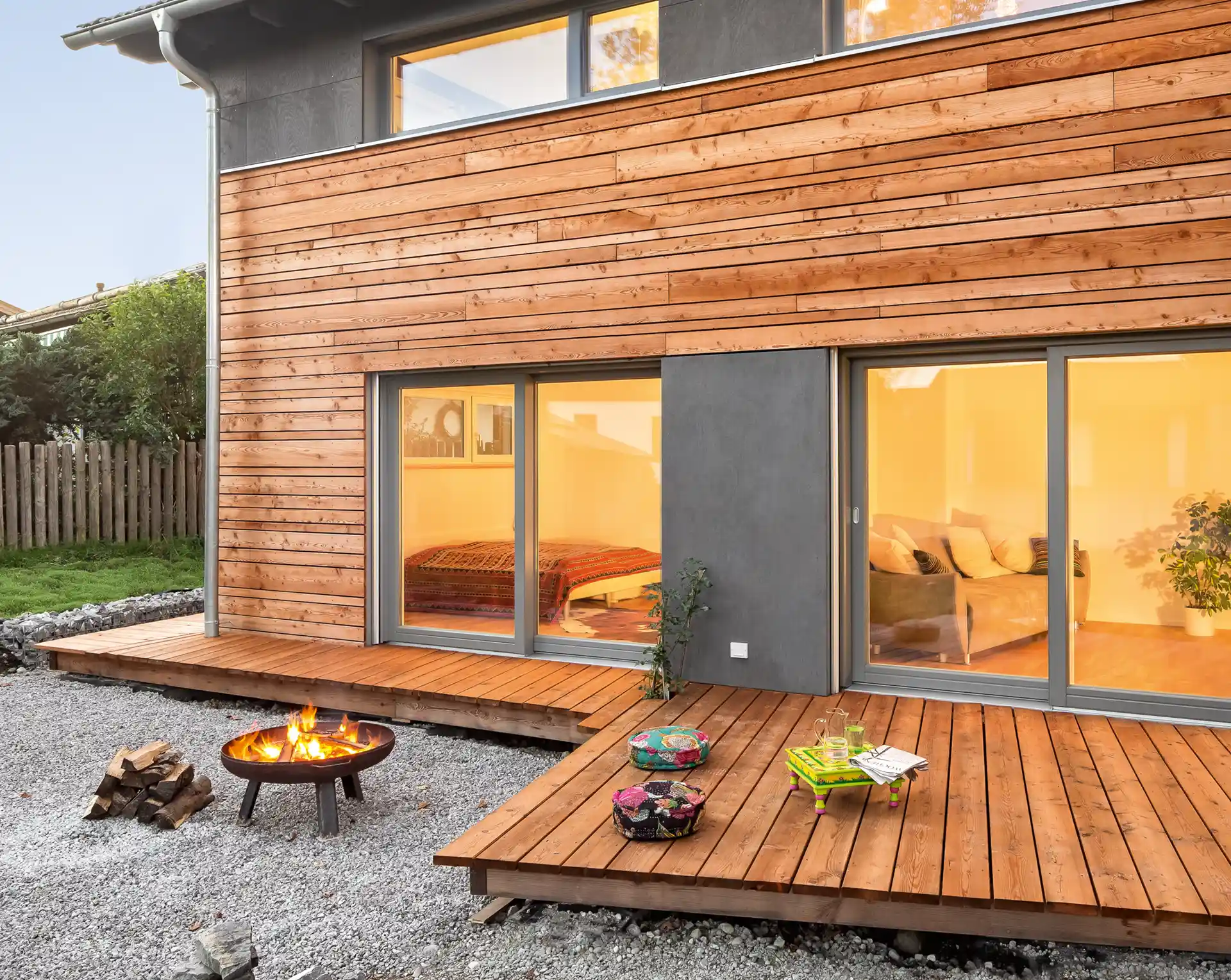 Modernes Holzhaus mit Holzdeck, Feuerschale, Kissen und einem Tisch im Freien mit Getränken im Abendlicht.
