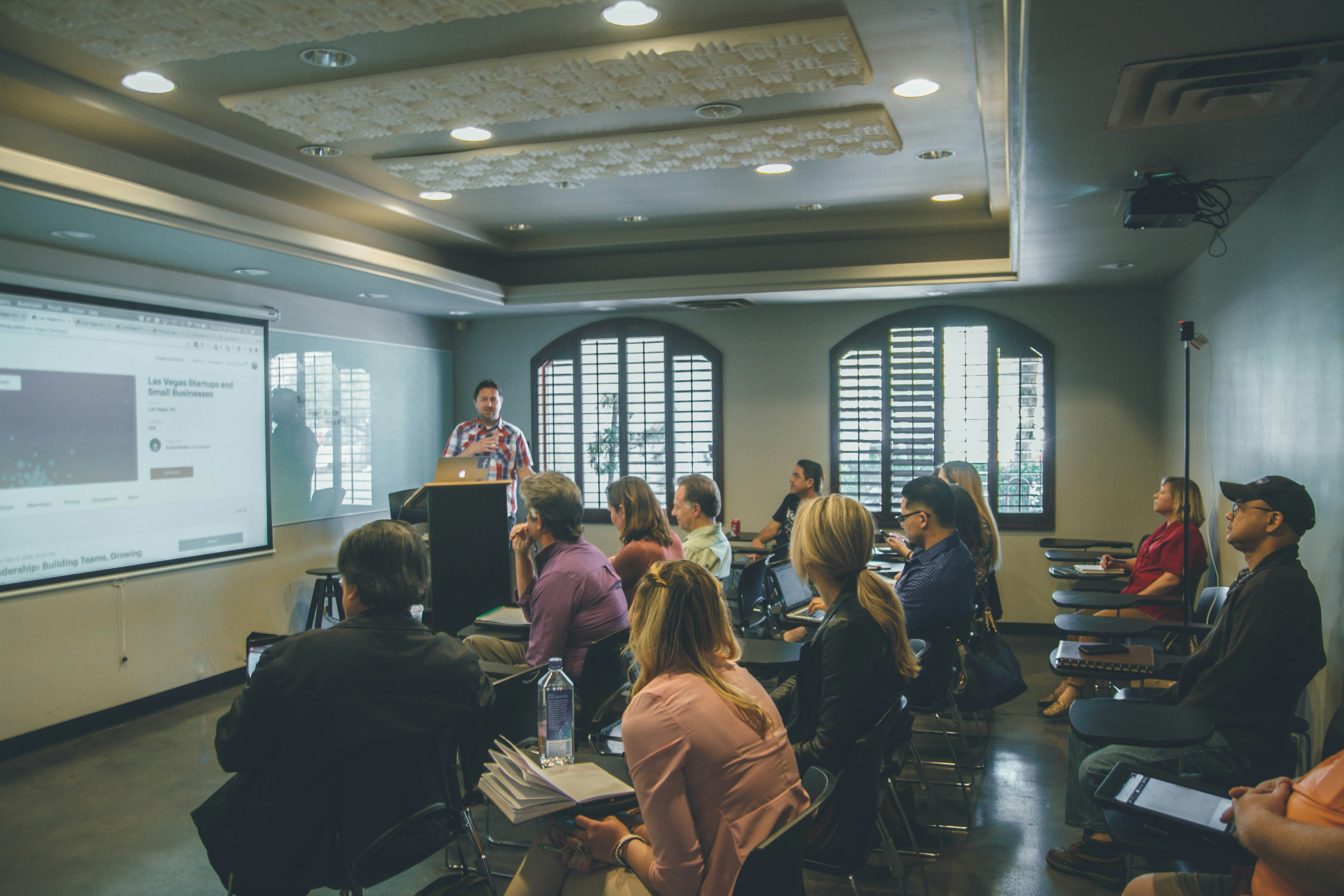 A class of students face a large screen on the left-hand wall. A teacher can be seen behind this at a podium. Behind him, two windows are on the far wall.