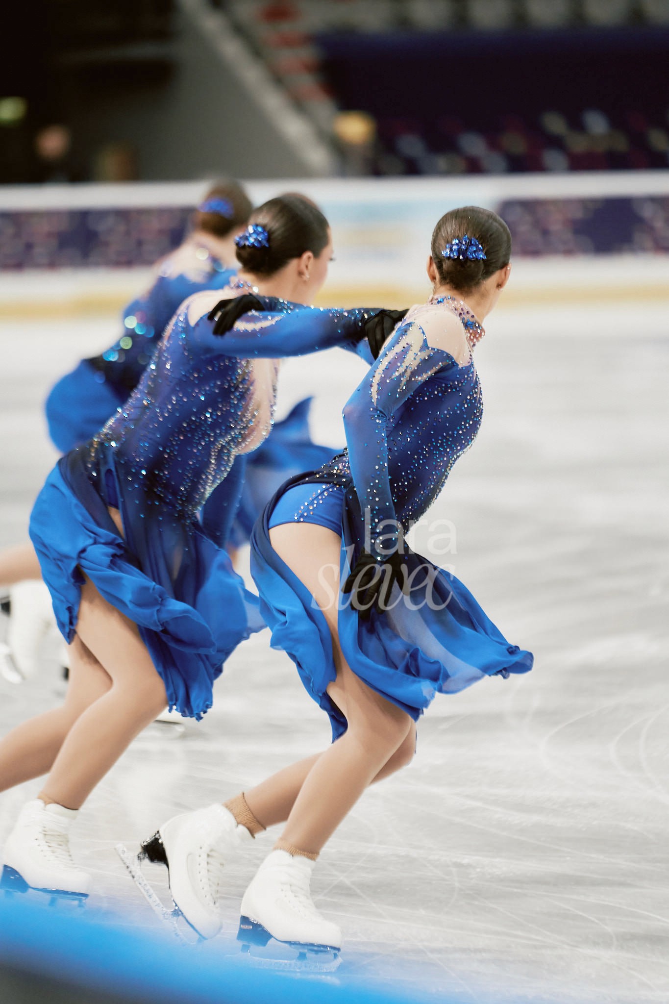 Drei Eiskunstläuferinnen im Synchroneiskunstlauf. In blauen, wehenden Kostümen. Team Haydenettes. ISU Synchronized Skating Championships 2026 in Salzburg. Fotografiert von Lara Slevec.