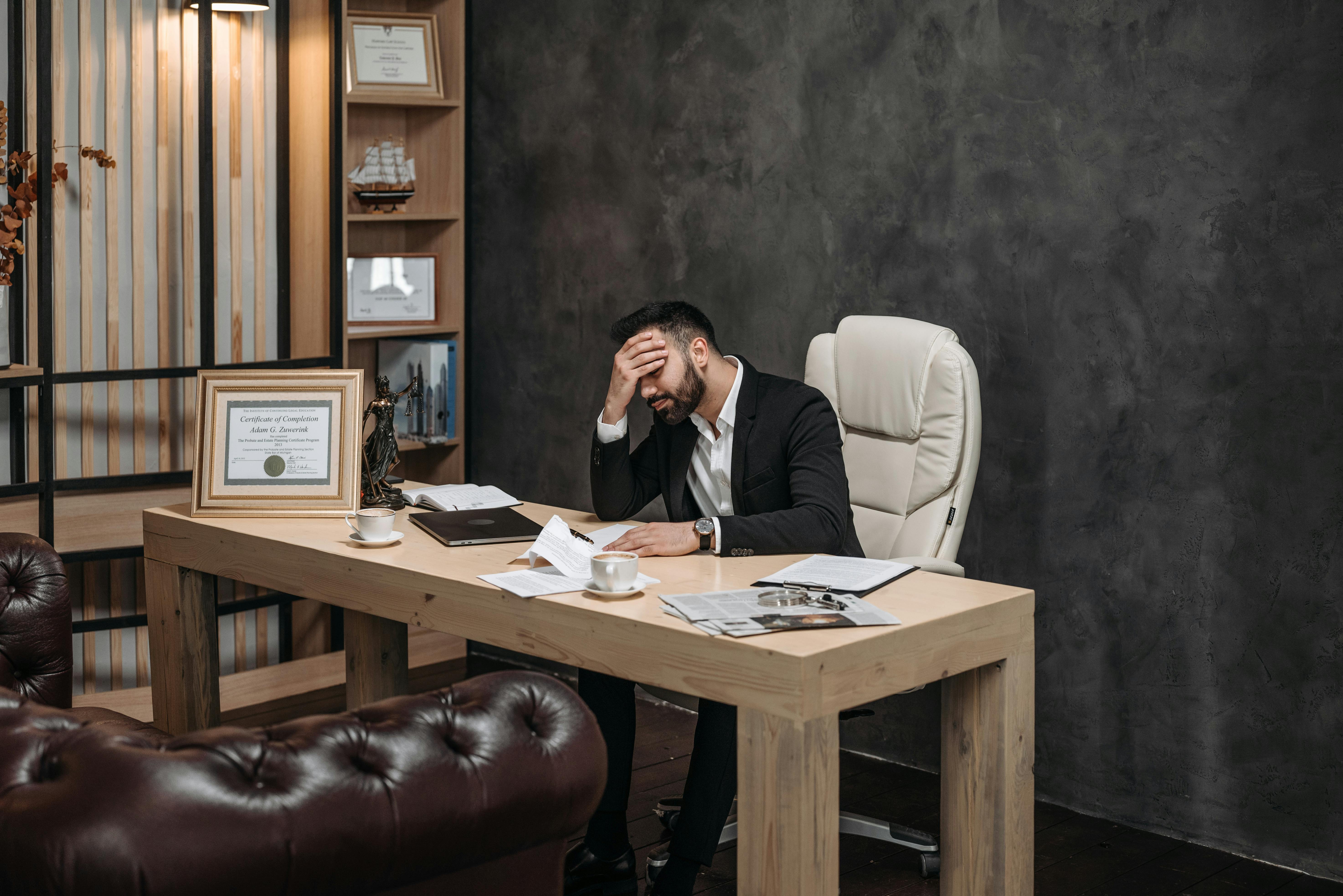 Man in Black Suit Sitting on His Office While Holding His Head