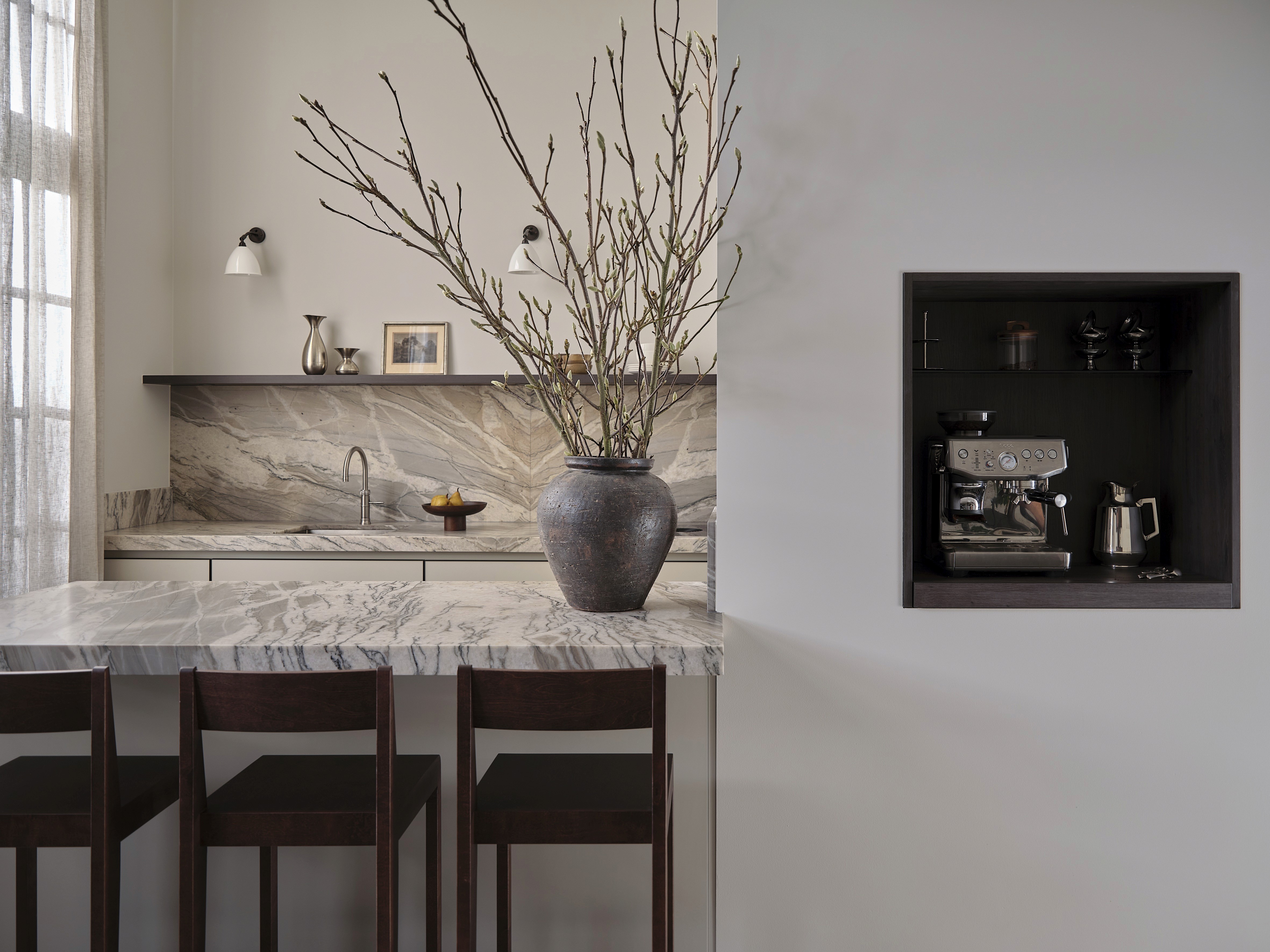 Kitchen with book-matched quartzite island and backsplash, ceramic vase with budding branches, built-in coffee station, and dark wood bar stools