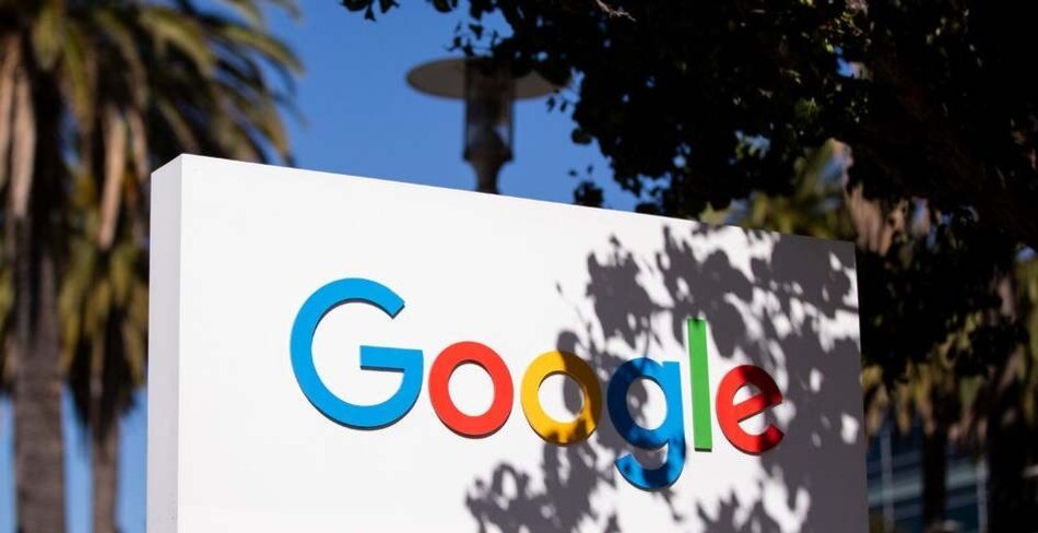 Google's colorful logo is displayed on a white sign, partially shaded by tree branches, in front of a palm-lined office building.