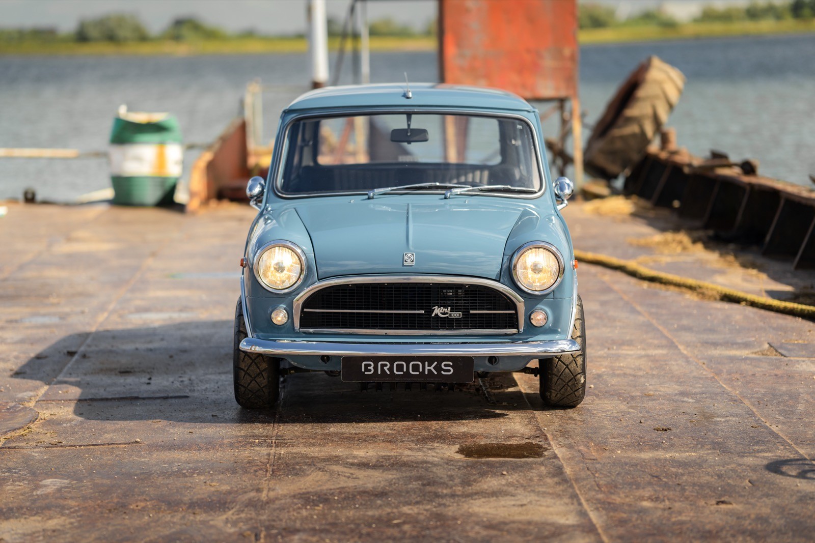 A light blue vintage car parked near a waterfront, with a cargo area and industrial backdrop.