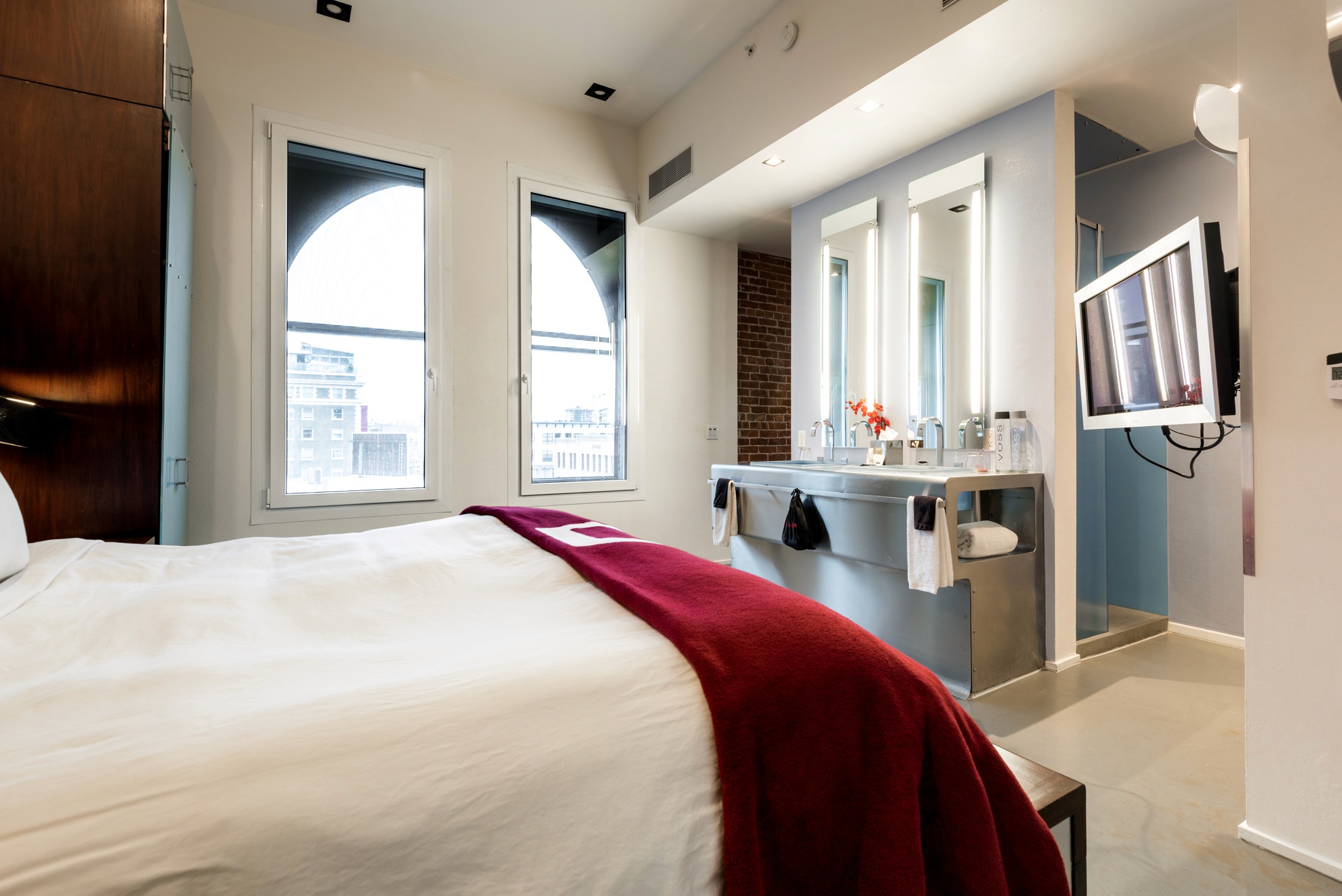 Bright modern hotel room featuring crisp white linens, deep red accents, and natural light streaming through large windows.