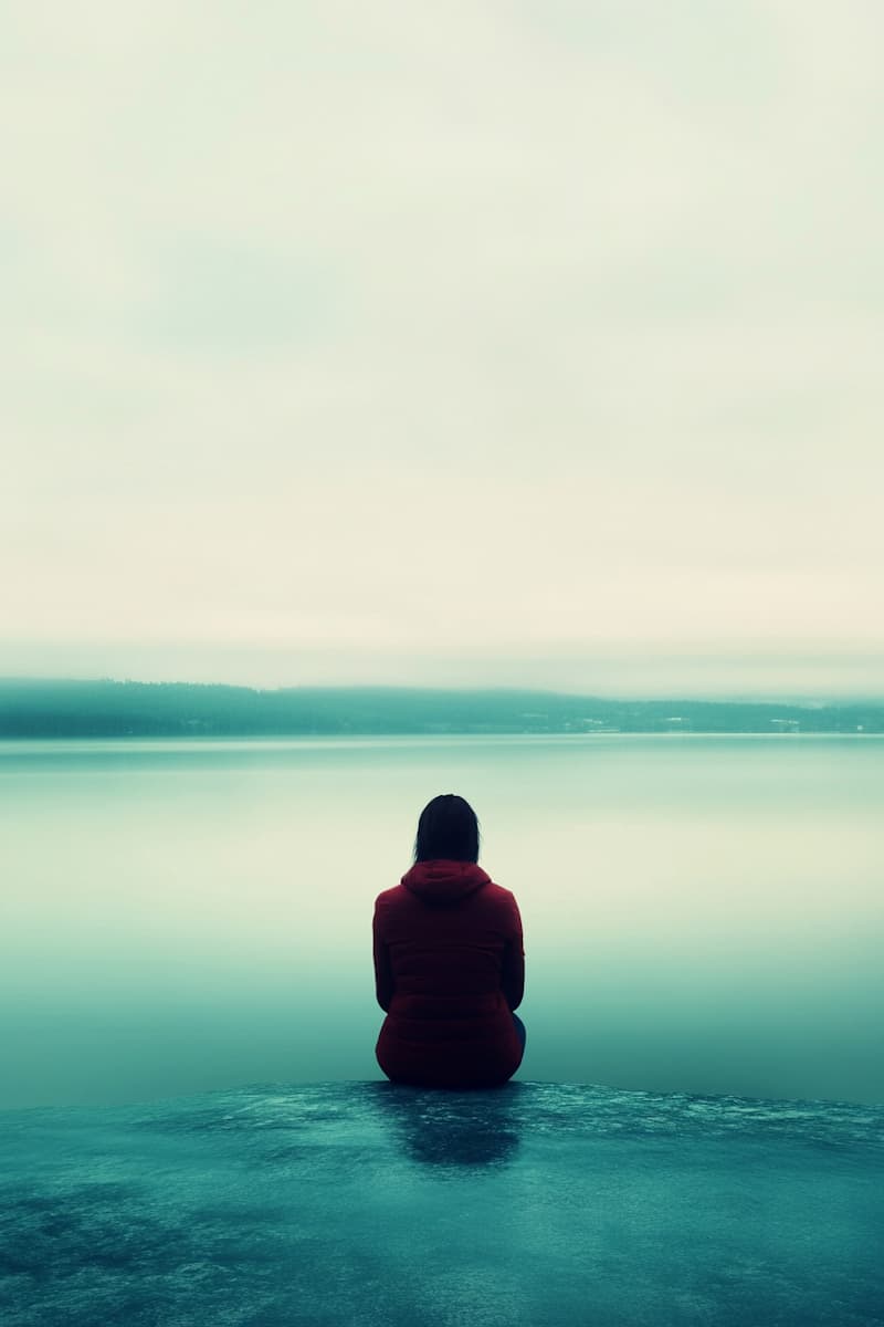 Einsamer Mann am Seeufer. Melancholische Stimmung, ruhiges Wasser und grauer Himmel.