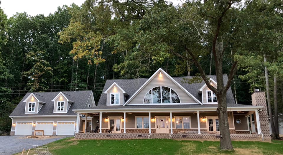 Front view of modern custom home with large front porch and three car garage with soft exterior lighting on at dusk with green grass front lawn with large tree.
