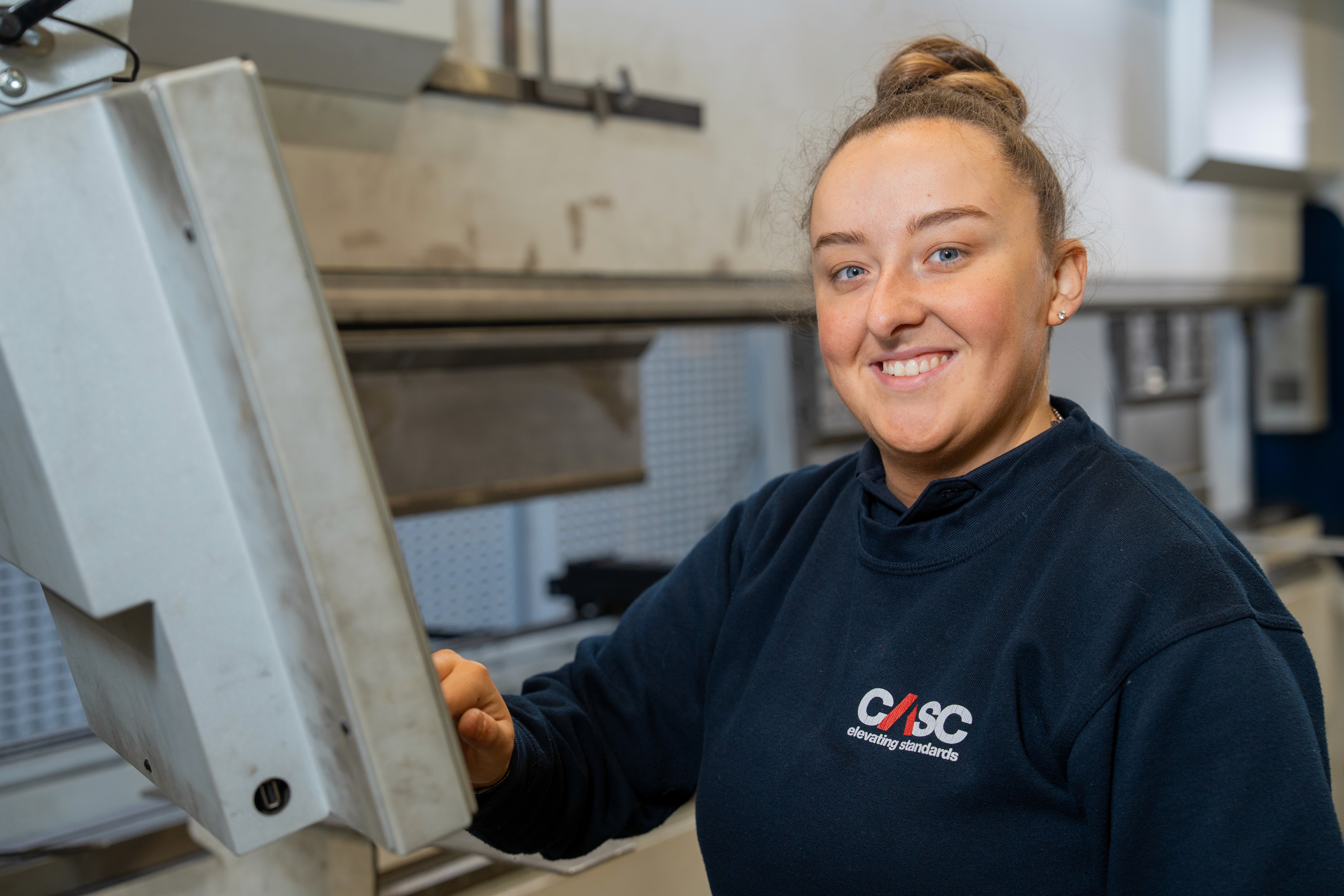 CASC team member in a factory environment reviewing a checklist and measuring a component as part of quality control and inspection work.