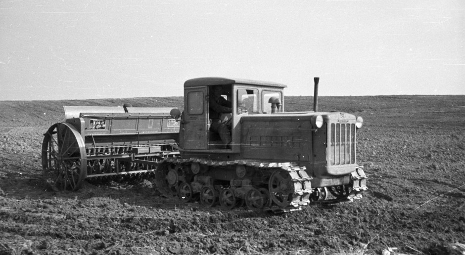 Two people in a workshop, one standing and smiling while the other leans over a car, working on it.