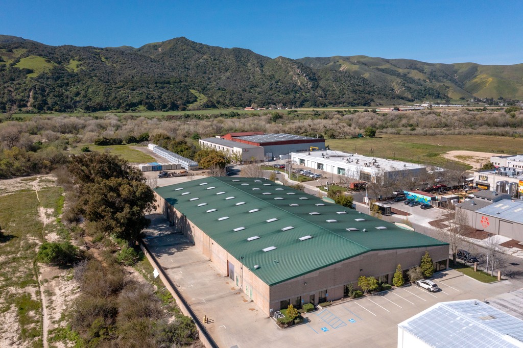 Wide aerial view of the same green-roofed warehouse facility from a higher angle, showing its relationship to adjacent light industrial buildings, a riparian corridor, and a ridgeline of tree-covered hills in the background.