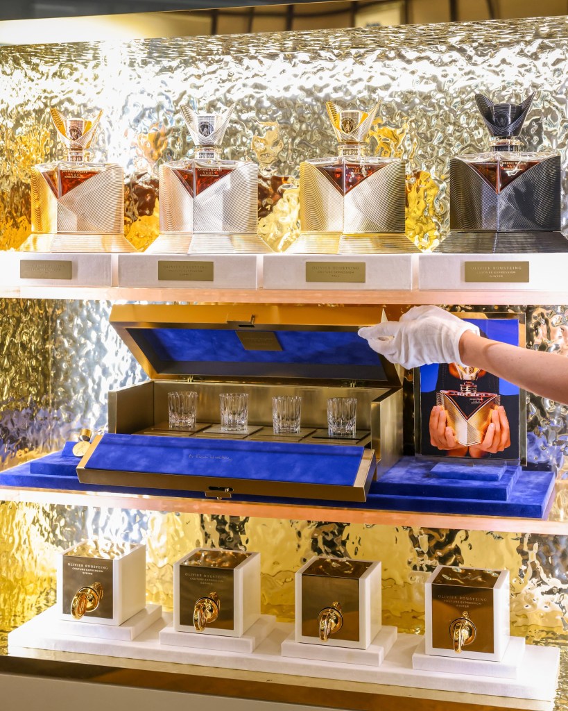 Display of Johnnie Walker Couture Blend decanters featuring sleek designs and a luxurious presentation, with a hand elegantly opening a box containing glasses against a golden backdrop.