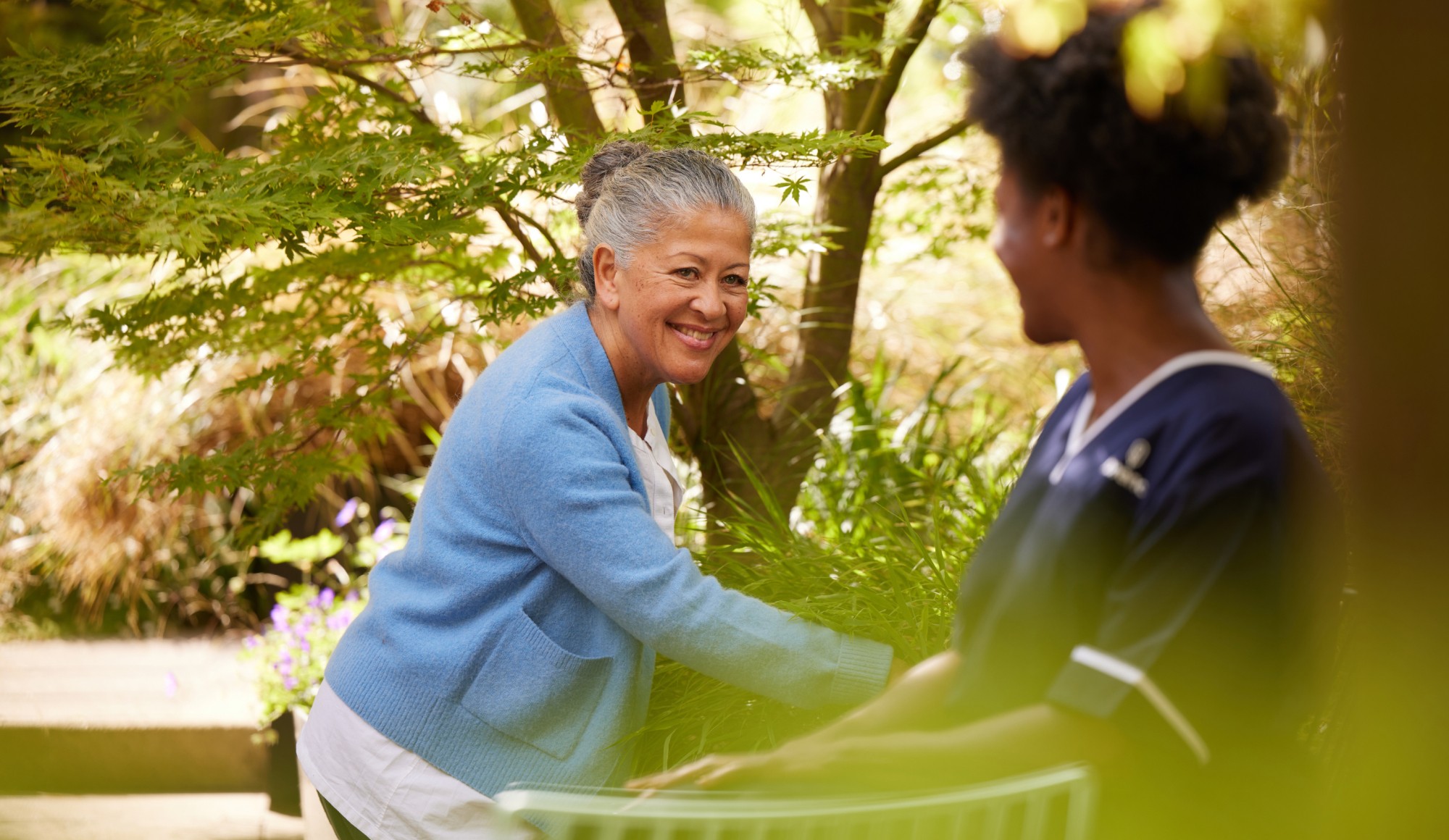 Older woman smiling and interacting with a Florence caregiver outdoors in a leafy garden setting.