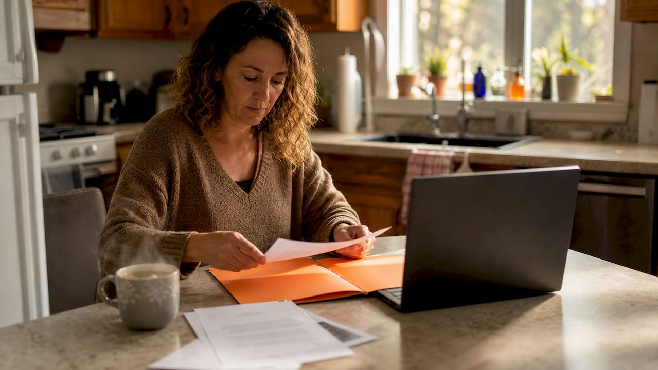 Parent reviewing grant application paperwork at a kitchen table with a laptop