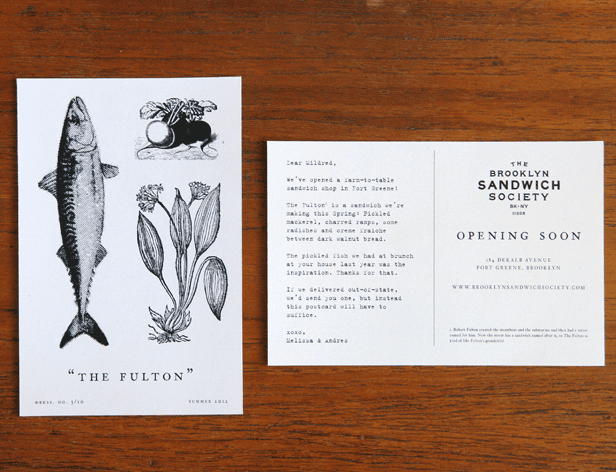 The image shows two pages from a book. The left page features a black and white illustration of a fish labeled "The Fulton," a plant illustration, and a small illustration of a root or bulb. The right page displays text that reads “Opening Soon,” followed by “The Brooklyn Sandwich Society,” “Opening Soon,” and “14 Dekals Avenue, Fort Greene, Brooklyn.” The text also mentions that the sandwich shop will open in Port Greene. The pages are placed on a wooden surface.