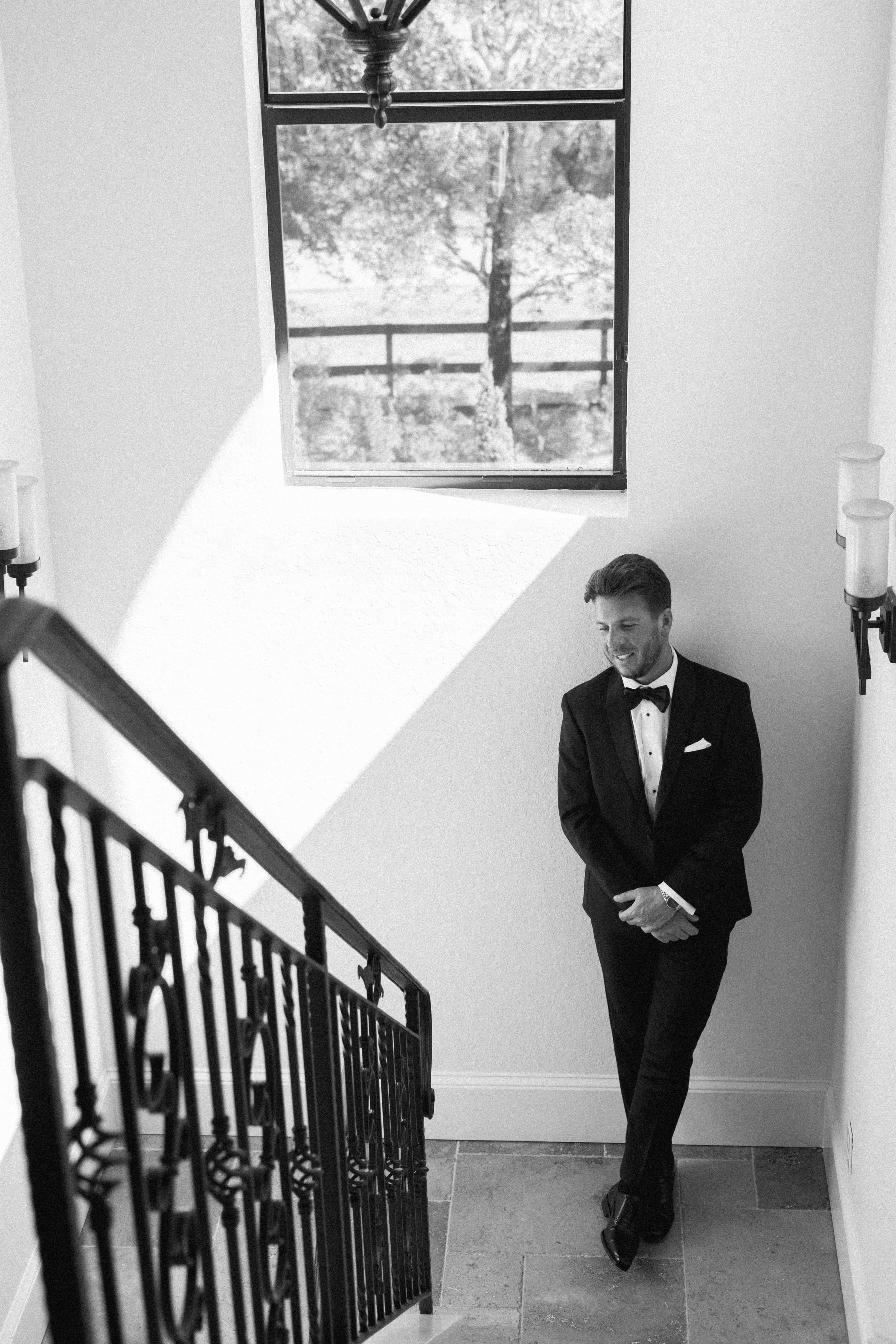 A black-and-white photo of a man in a tuxedo standing on a staircase, looking down at his feet. Sunlight streams in through a large window.