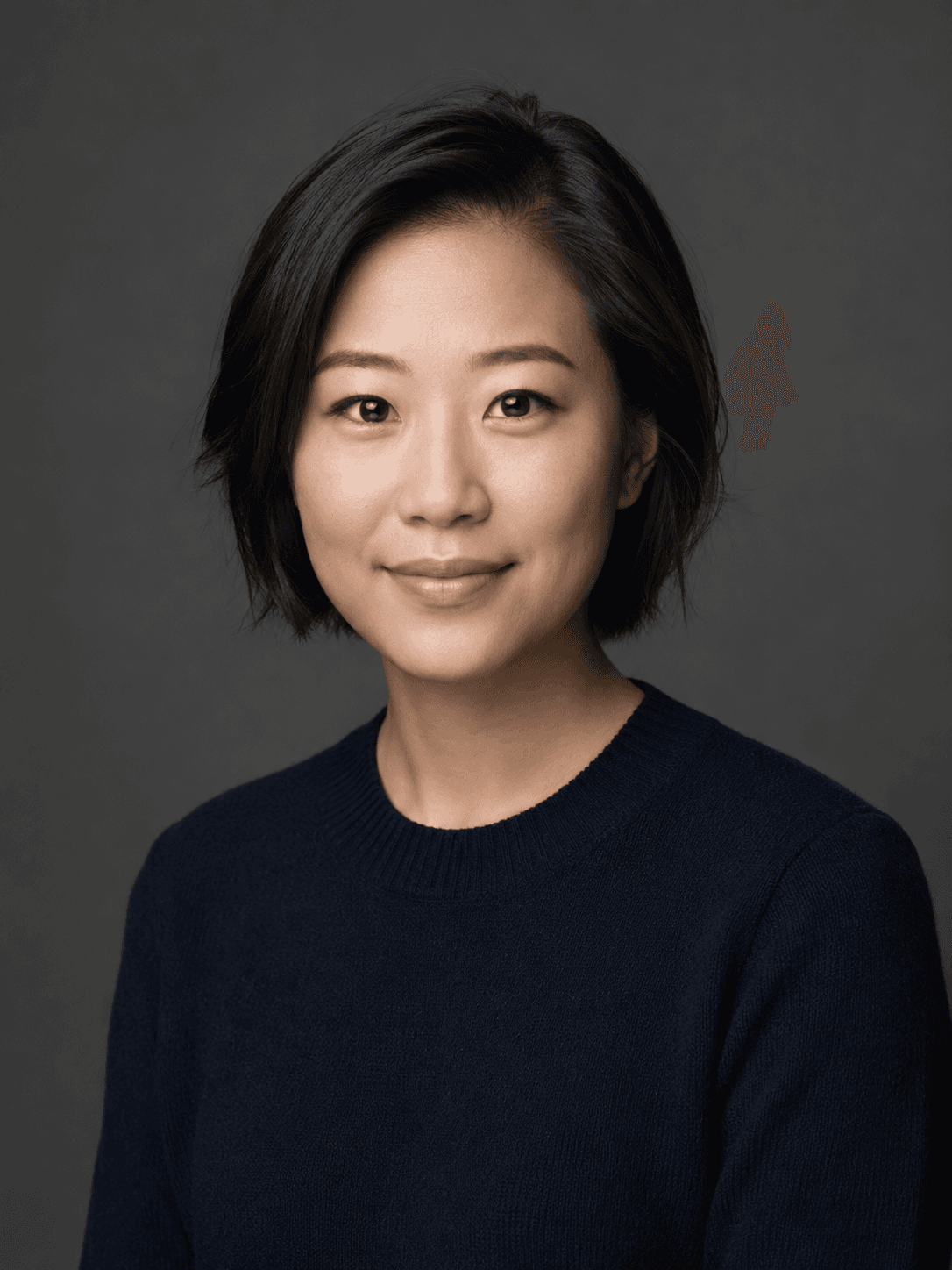A smiling person with short dark hair, wearing a navy sweater, against a gray background.