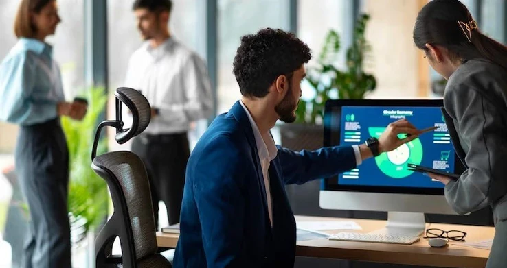 Homem apresentando um painel de indicadores num monitor para colegas num escritório