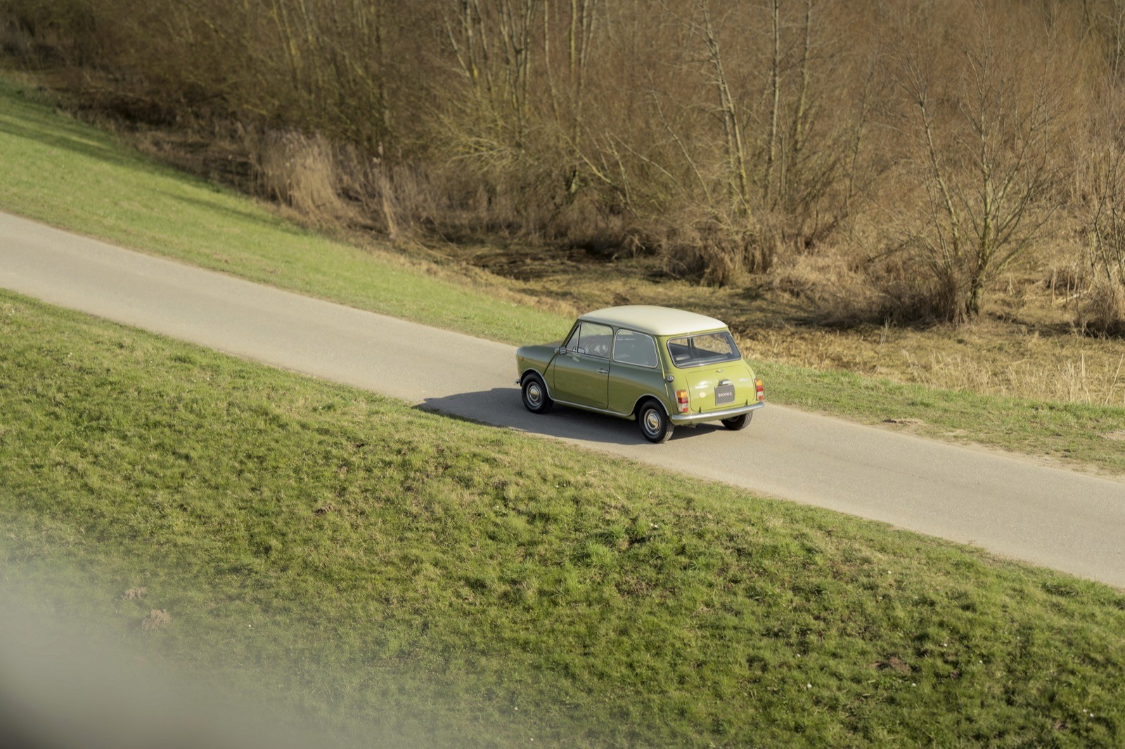 A light green car driving on a rural road surrounded by grass and trees in the background.