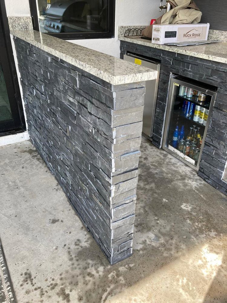 Outdoor kitchen with a stone tile wall, granite countertop, and built-in refrigerator.