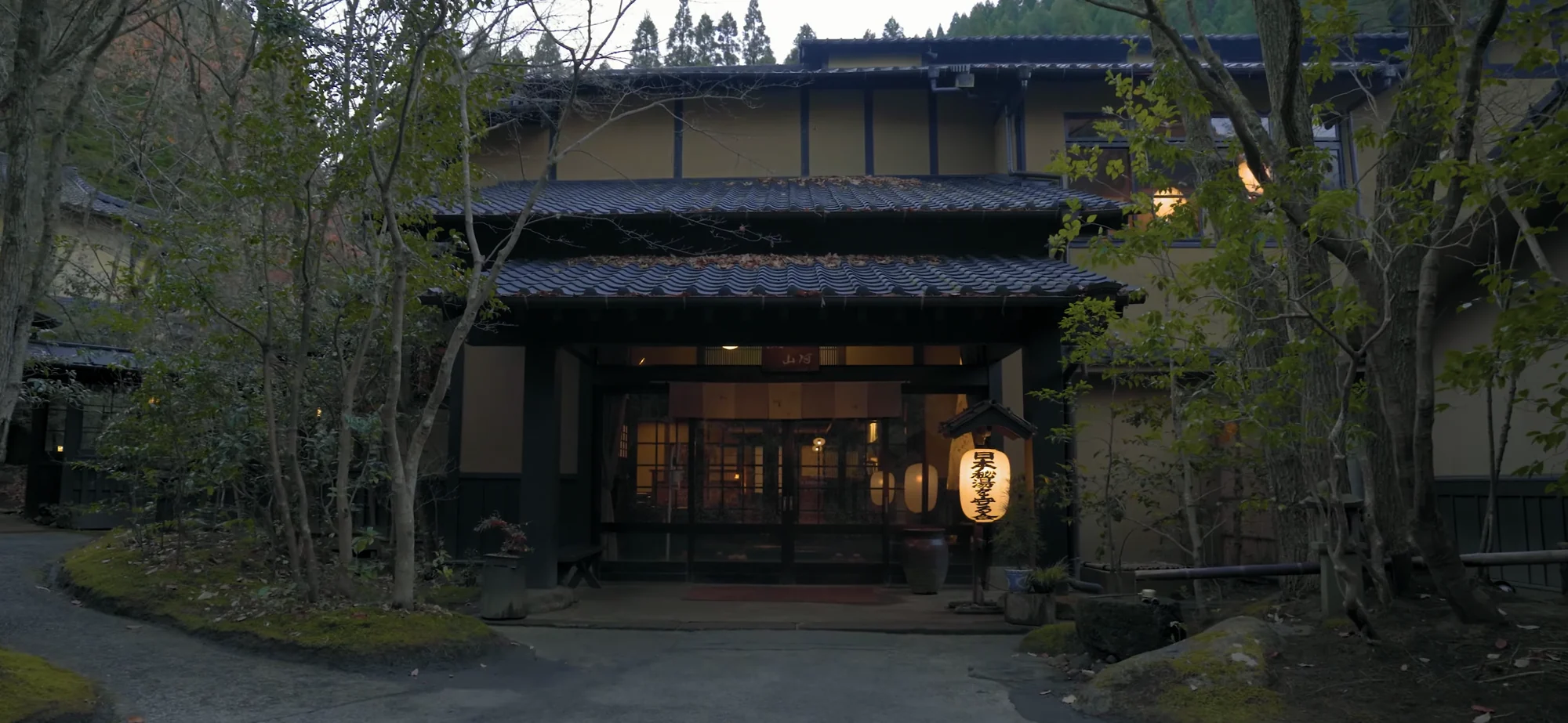 A dark timber ryokan frontage with a lantern burning at the entrance at dusk