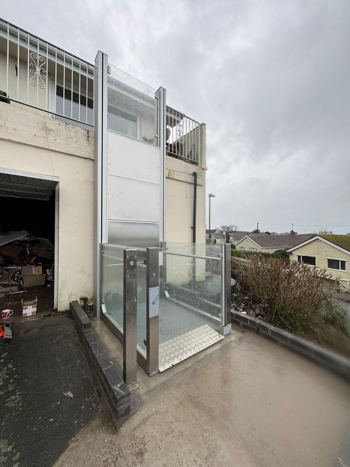 Outdoor vertical platform step lift raised to upper balcony level at a coastal residential property — full stainless steel column and glazed platform enclosure visible against rendered property wall