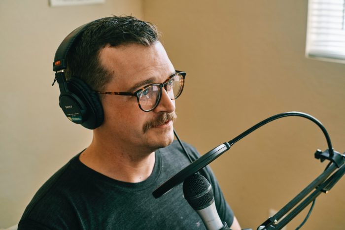 Man wearing glasses and headphones speaking into a microphone in a home podcast setup.