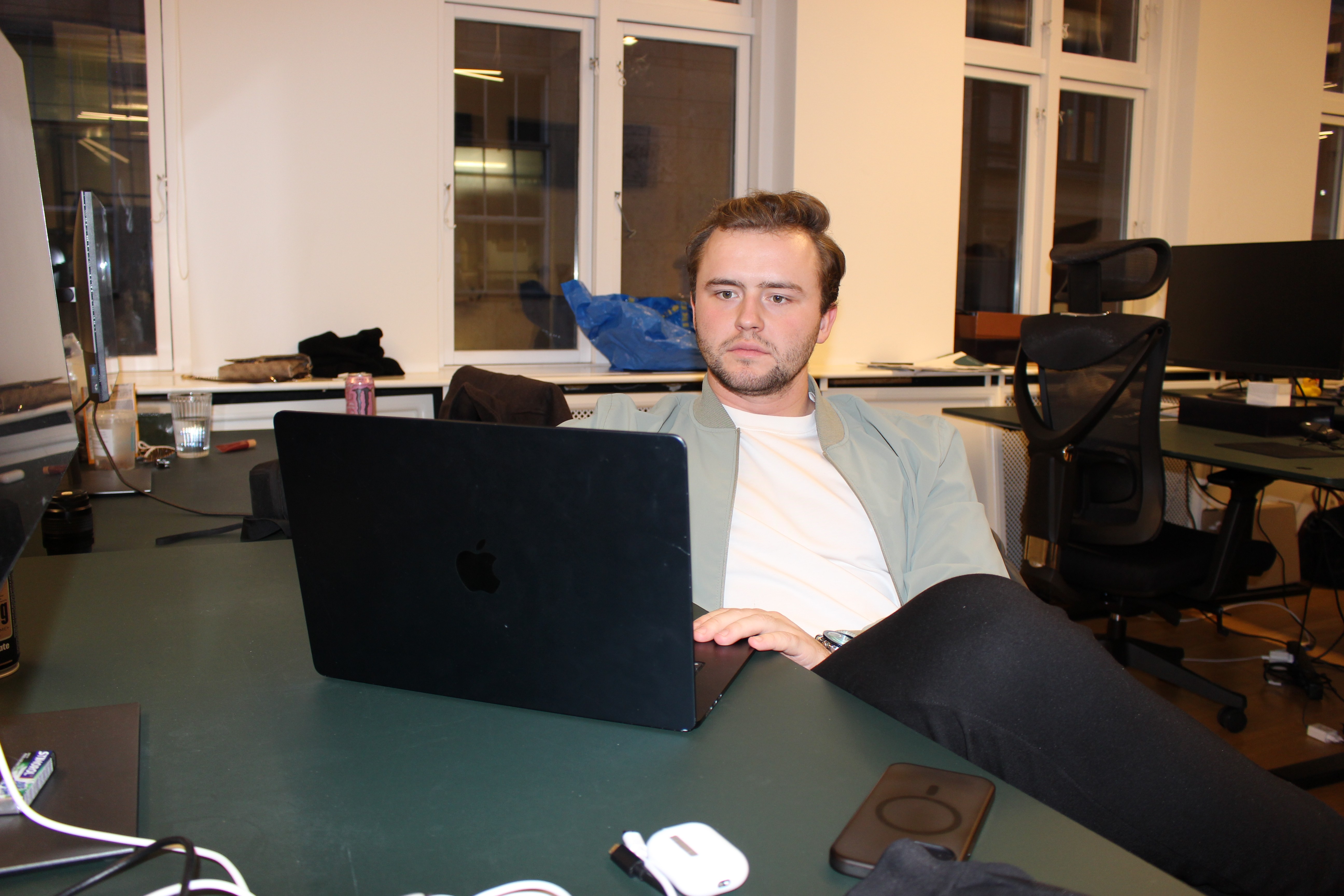 Two people at a wooden desk with a laptop and coffee, chatting and laughing by a large window