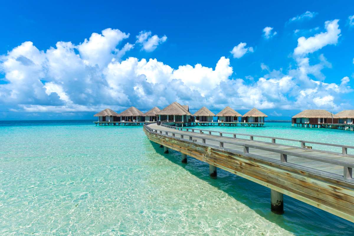A wooden pier leads toward several thatched-roof bungalows over turquoise water under a blue sky with white clouds.