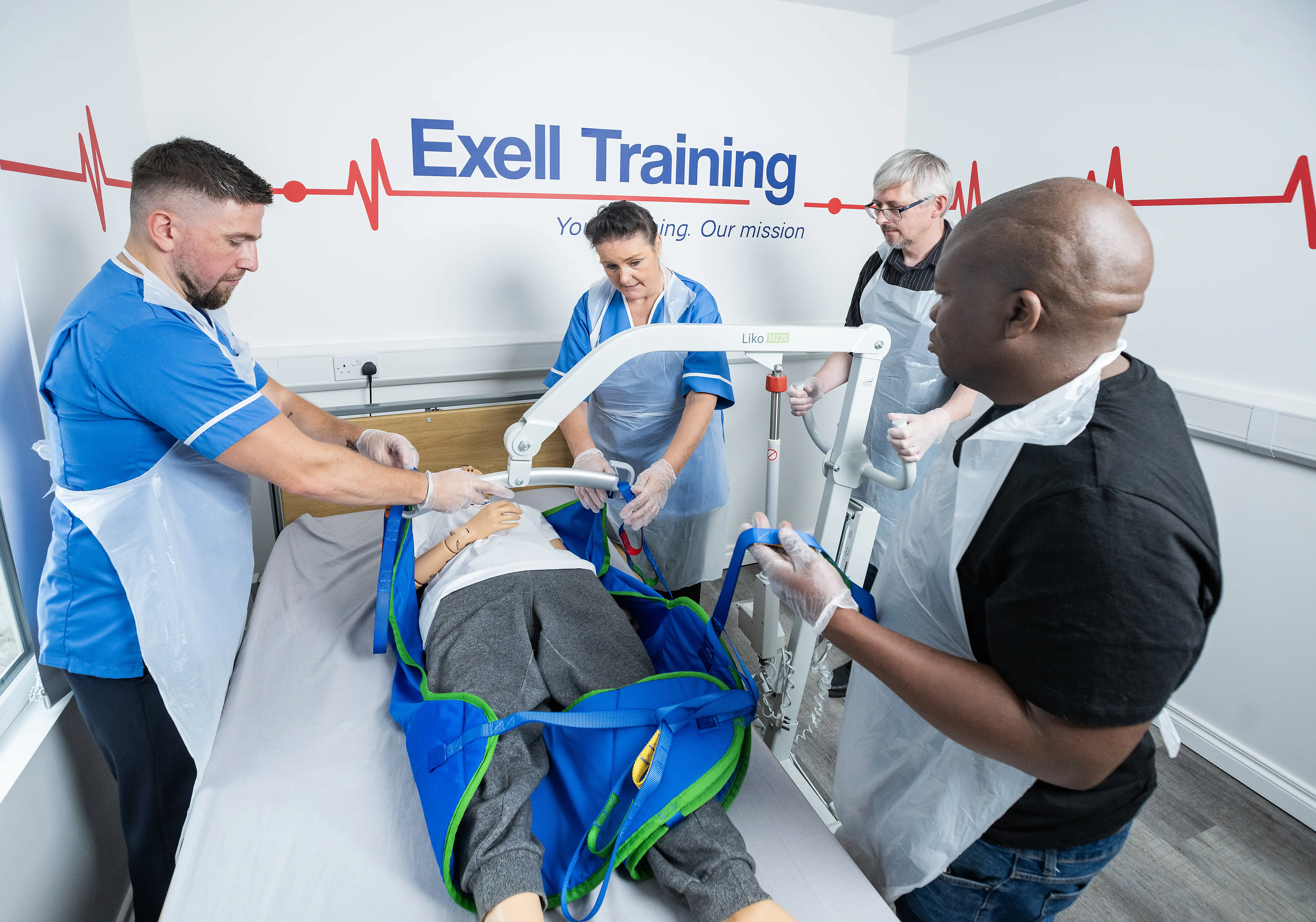 Group of healthcare trainees at Exell Training using a patient hoist to practise safe manual handling techniques on a medical mannequin.