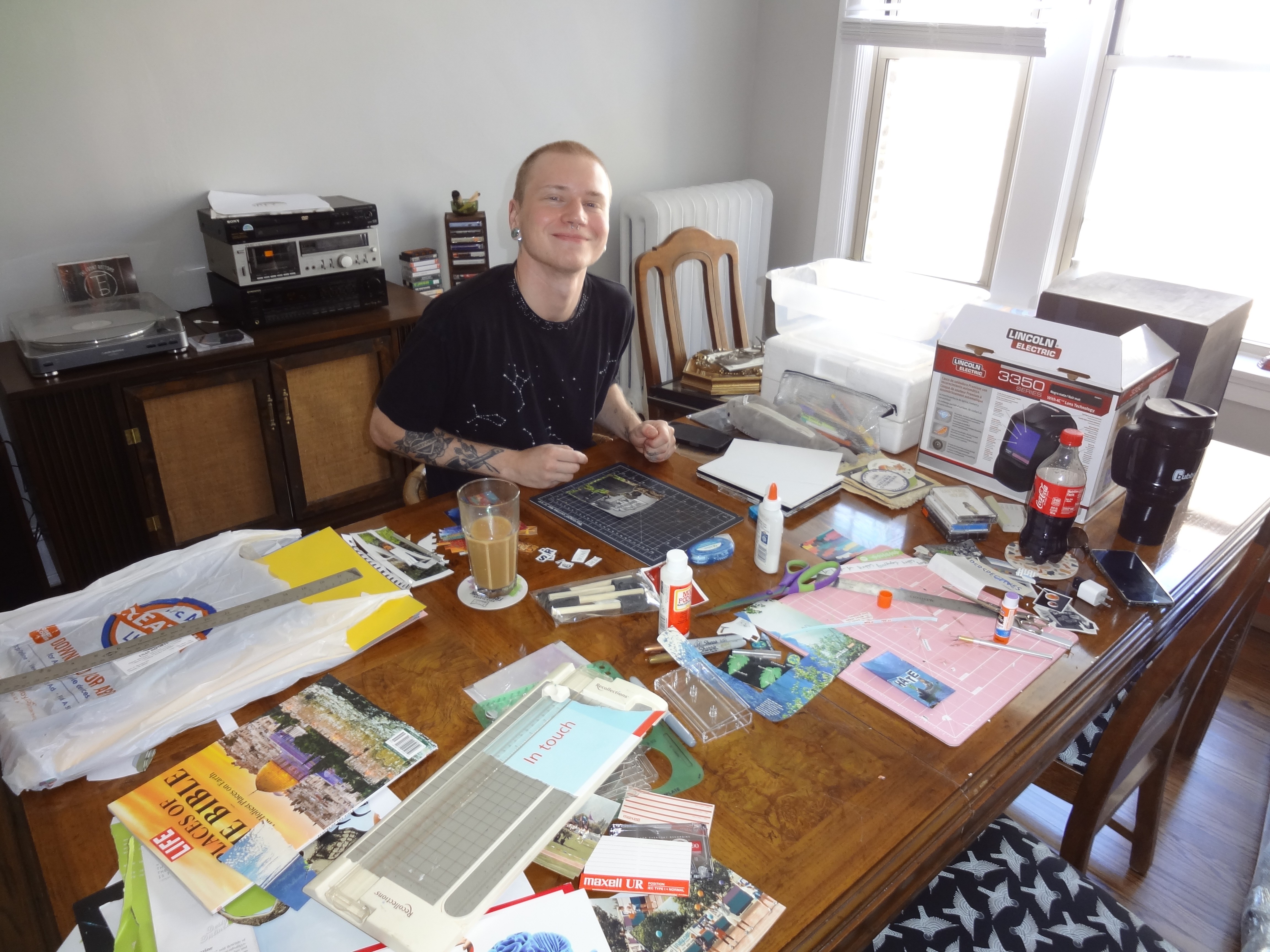 my roommate sitting at the table , which is covered in craft supplies for collage
