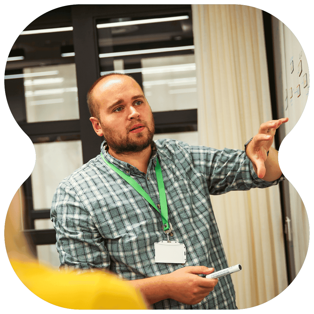 Person seated at a table with coloured markers in front of them, wearing a patterned shirt and leaning forward with hands clasped.