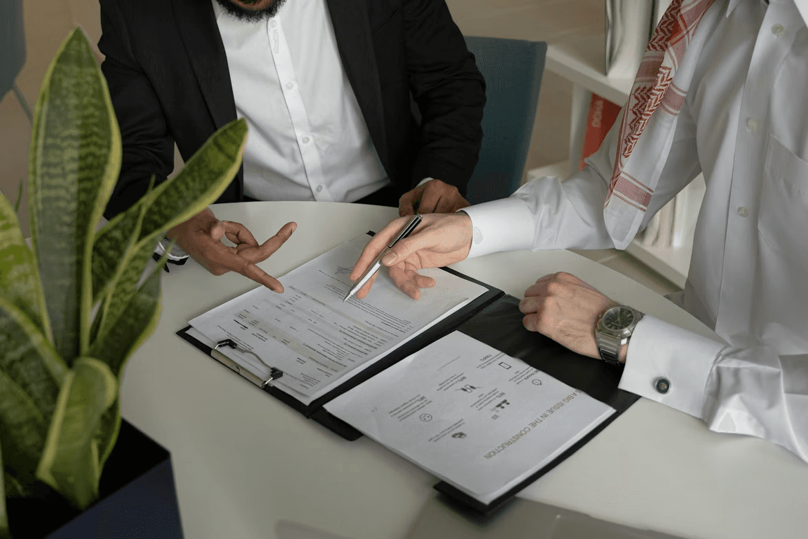 Two people sit at a table reviewing paperwork on a clipboard, with one person pointing at specific lines using a pen while the other gestures toward the form.
