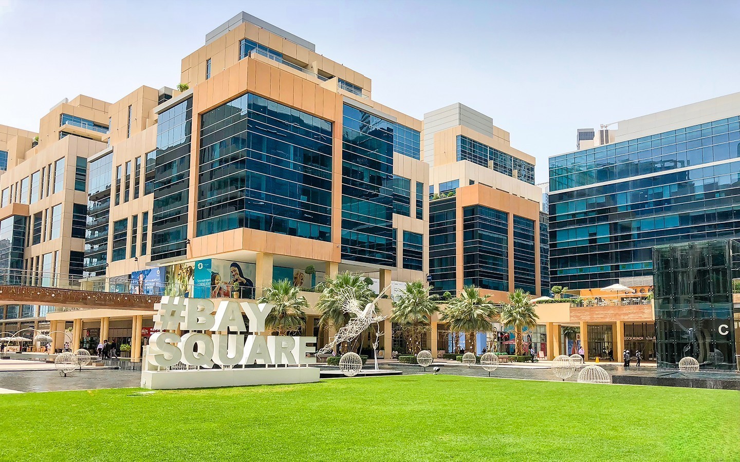 Modern glass buildings and green lawn at Bay Square in Business Bay with public art and walkways.