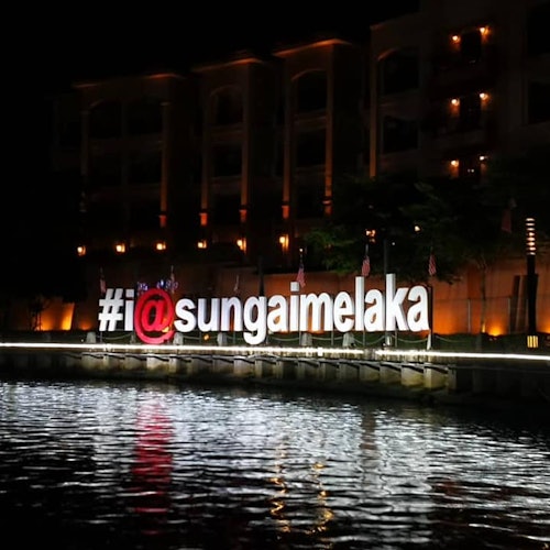 The large illuminated sign "#i@sungaimelaka" is set against the waterfront at night, with a building lit in the background.
